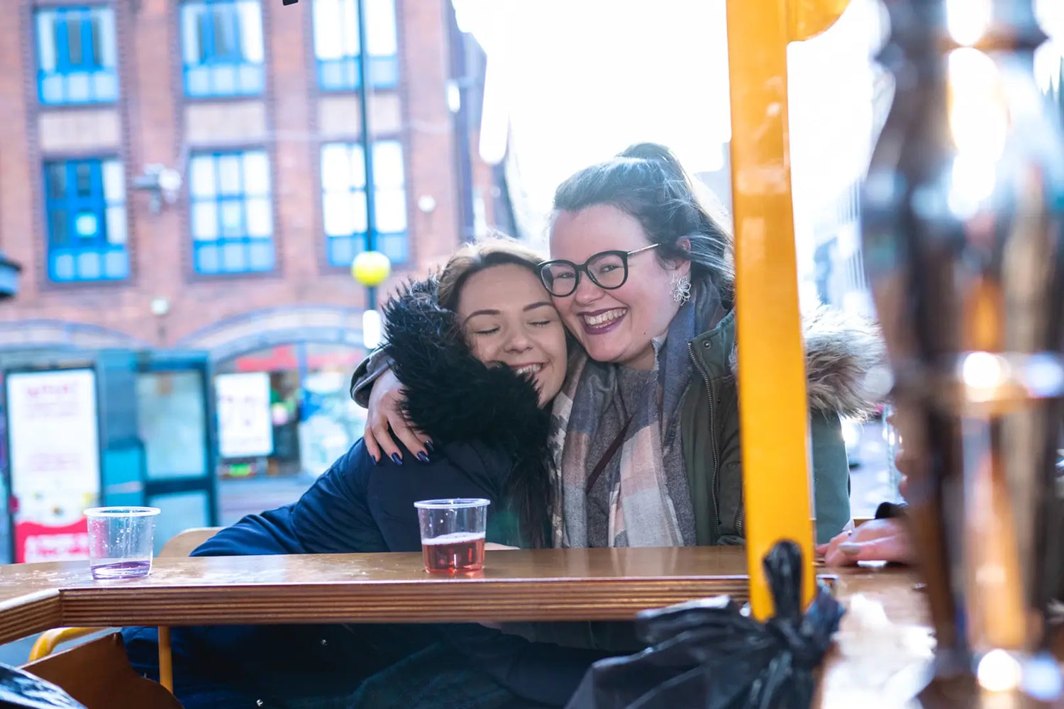 Two women embrace warmly at a bar on a bustling city street, celebrating with Wee Toast Tours.