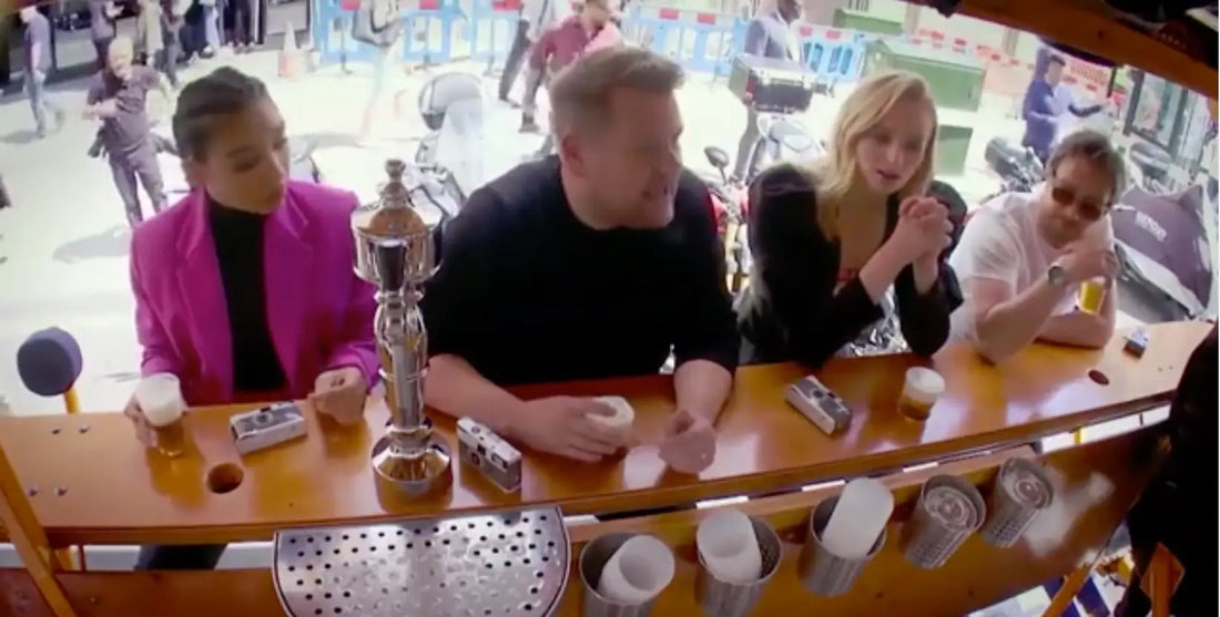 A group of people enjoying drinks at a bar, with James Corden and a bartender serving them - Wee Toast Tours experience.