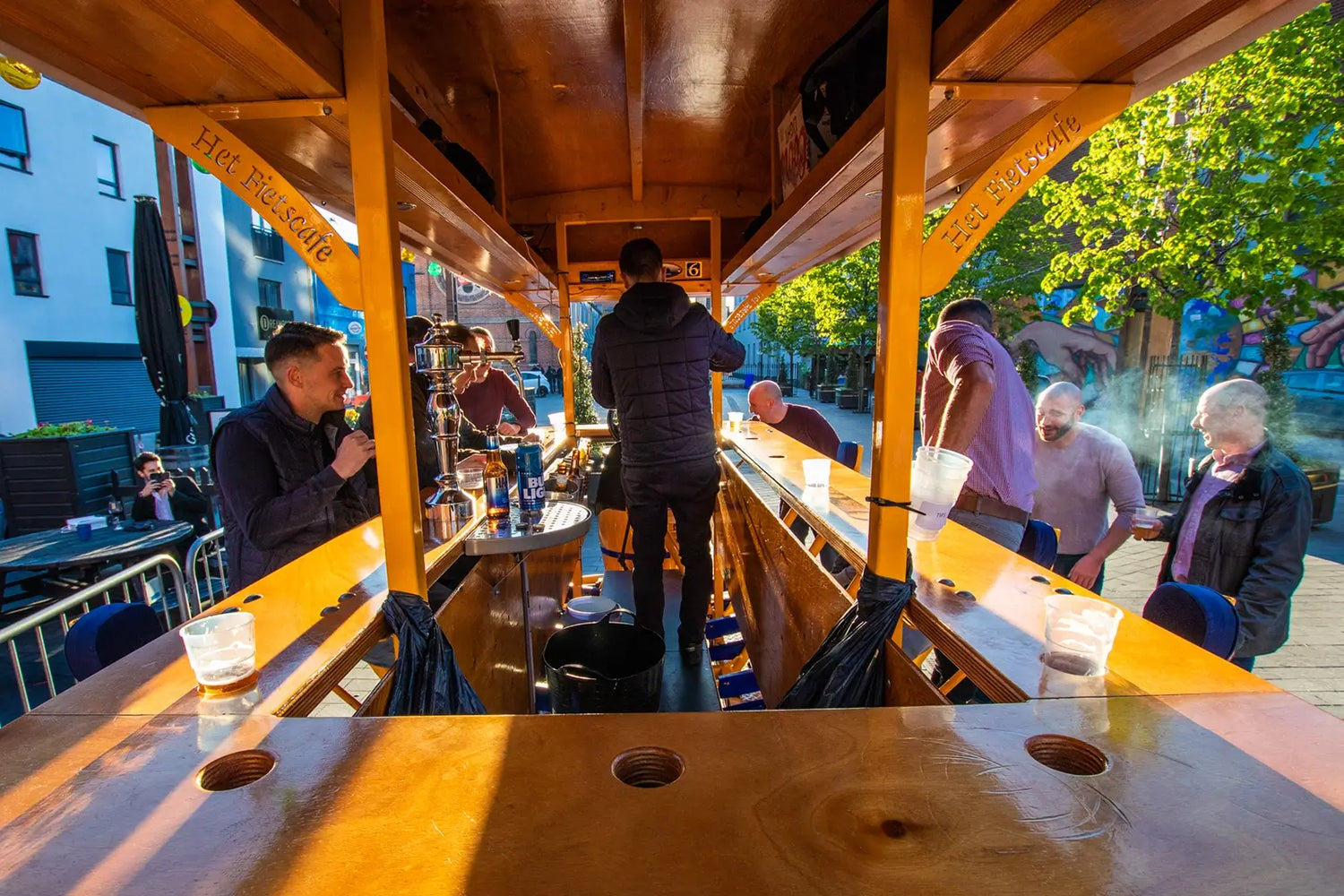 A vibrant outdoor bar setup, with patrons enjoying drinks at wooden counters under sunlight, surrounded by greenery and urban decor.