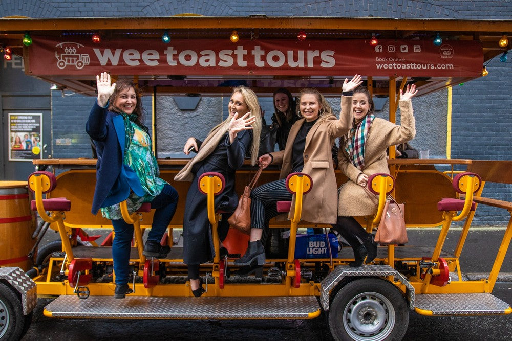 Four women pose on a bike with a sign for Wee Toast Tours, smiling and enjoying their outing.